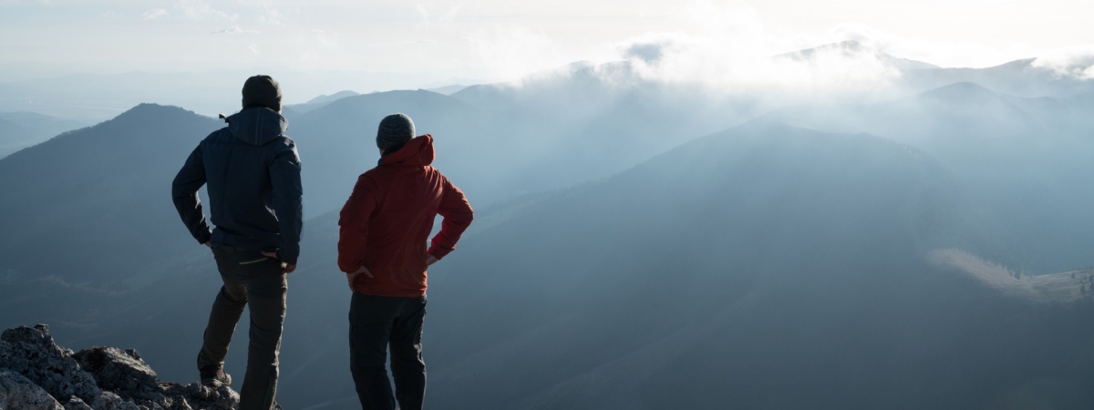 Two men standing standing with trekking poles on cliff edge and looking at sunset rays over the clouds. Successful summit concept image.